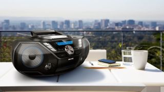 The PHILIPS AZB798T boombox on a table outdoors, beside a coffee cup and glass. In the background there's a view of a city.