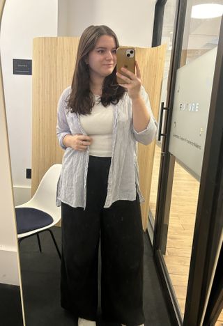 editor brooke knappenberger stands in front of a mirror wearing a button down shirt and black pants