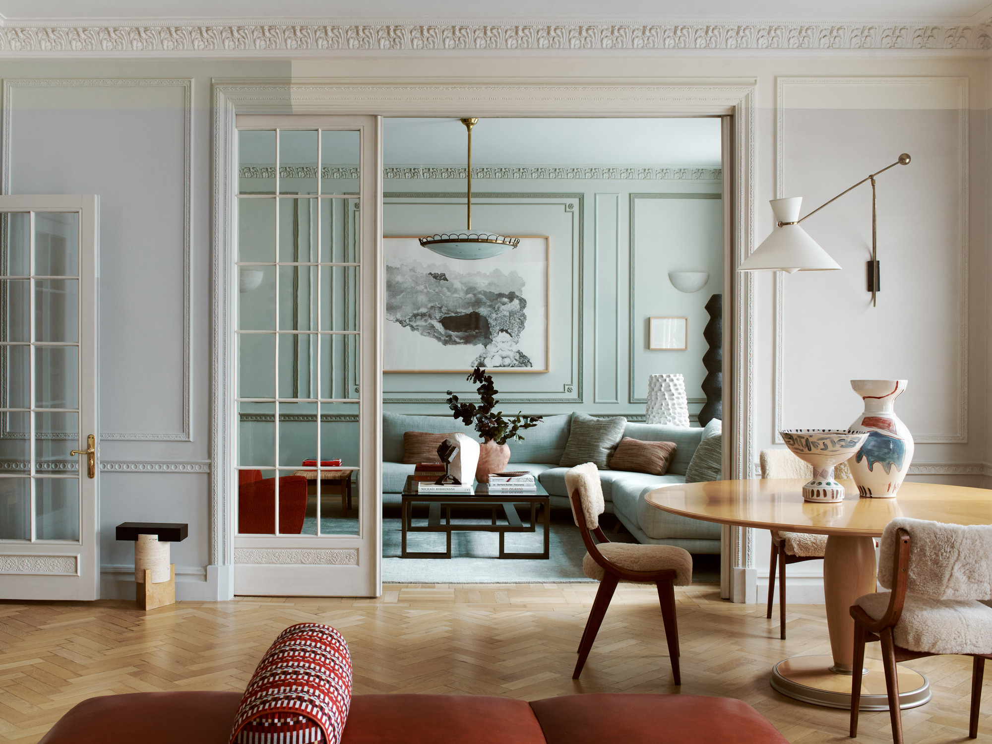 Image of a traditional living room that has glass doors that open into a smaller living room. There is crown molding and trim on the walls that are painted in different colors. There is a round dining table in the first room.