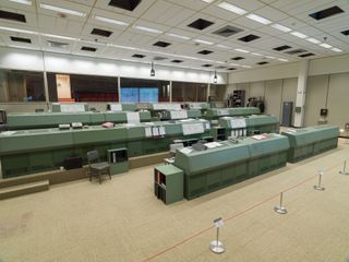 Alternative interior view of the recreated Johnson Space Centre in Houston featuring light coloured flooring, green workstations, shelving units and rails with different coloured jackets