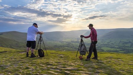 Two men on top of a hill in the Peak District stood next to their tripods and shooting towards the sunset over an impressive landscape