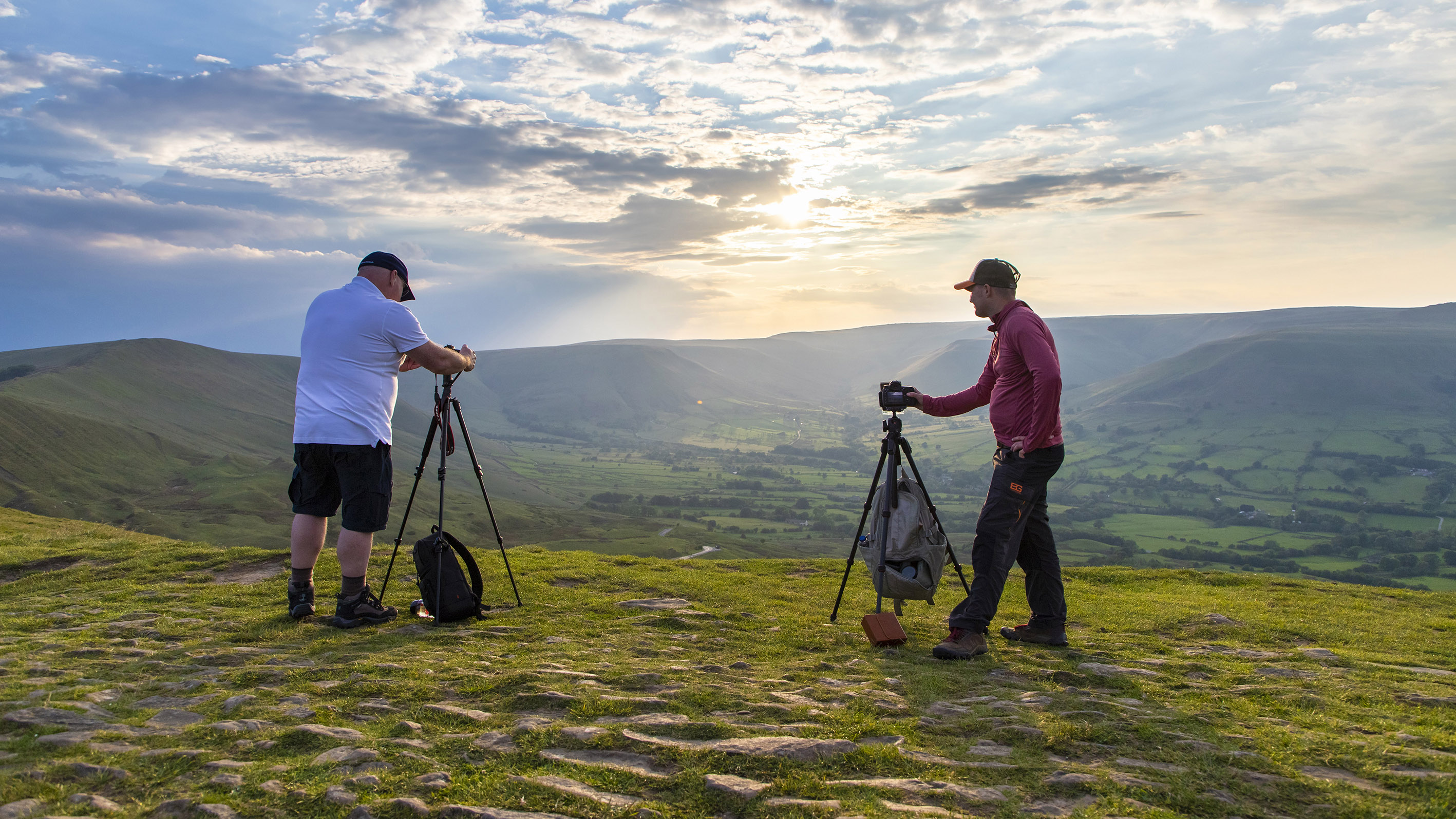 Two men on top of a hill in the Peak District stood next to their tripods and shooting towards the sunset over an impressive landscape