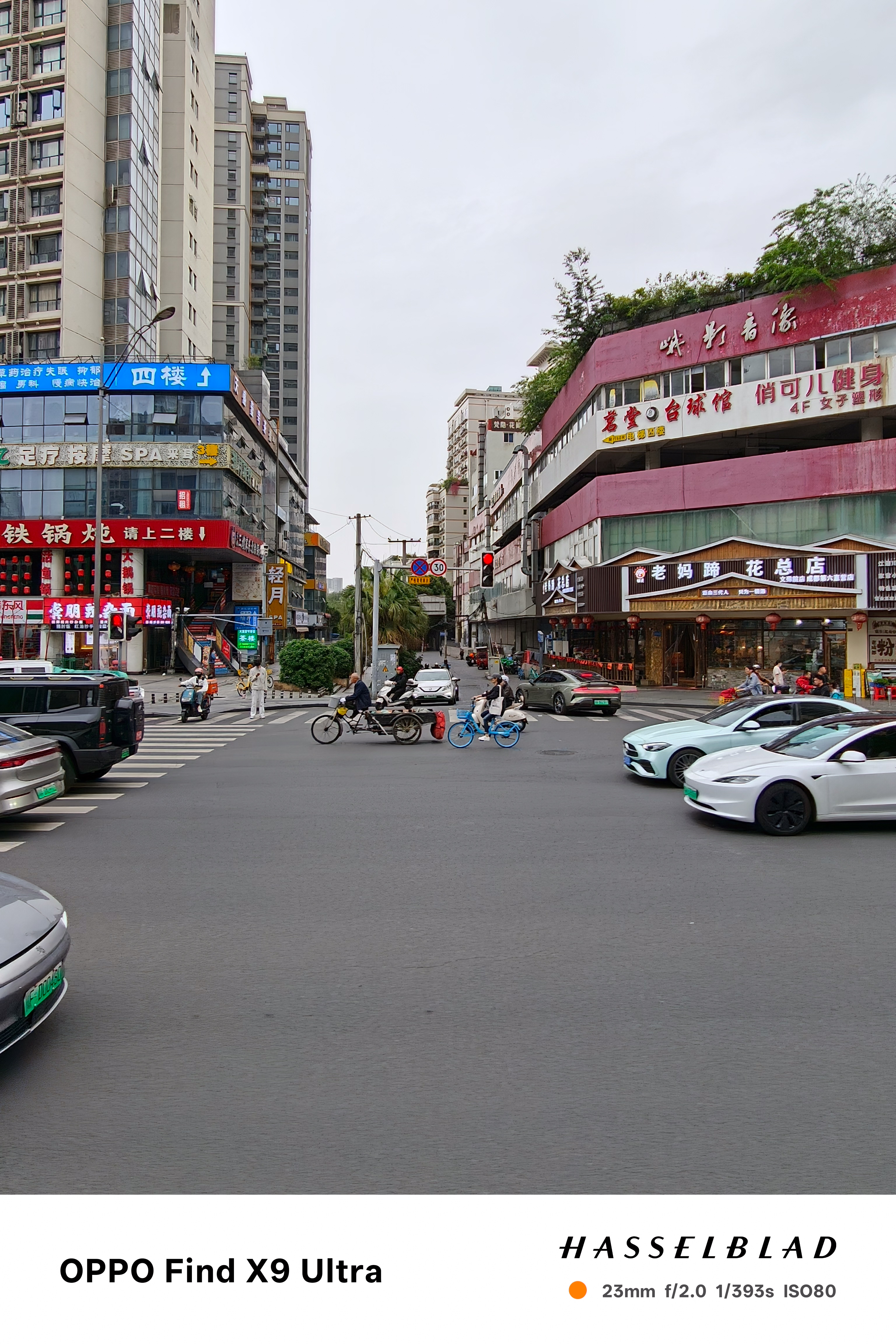 Busy city intersection with scooters, cars, and shopfronts under gray skies