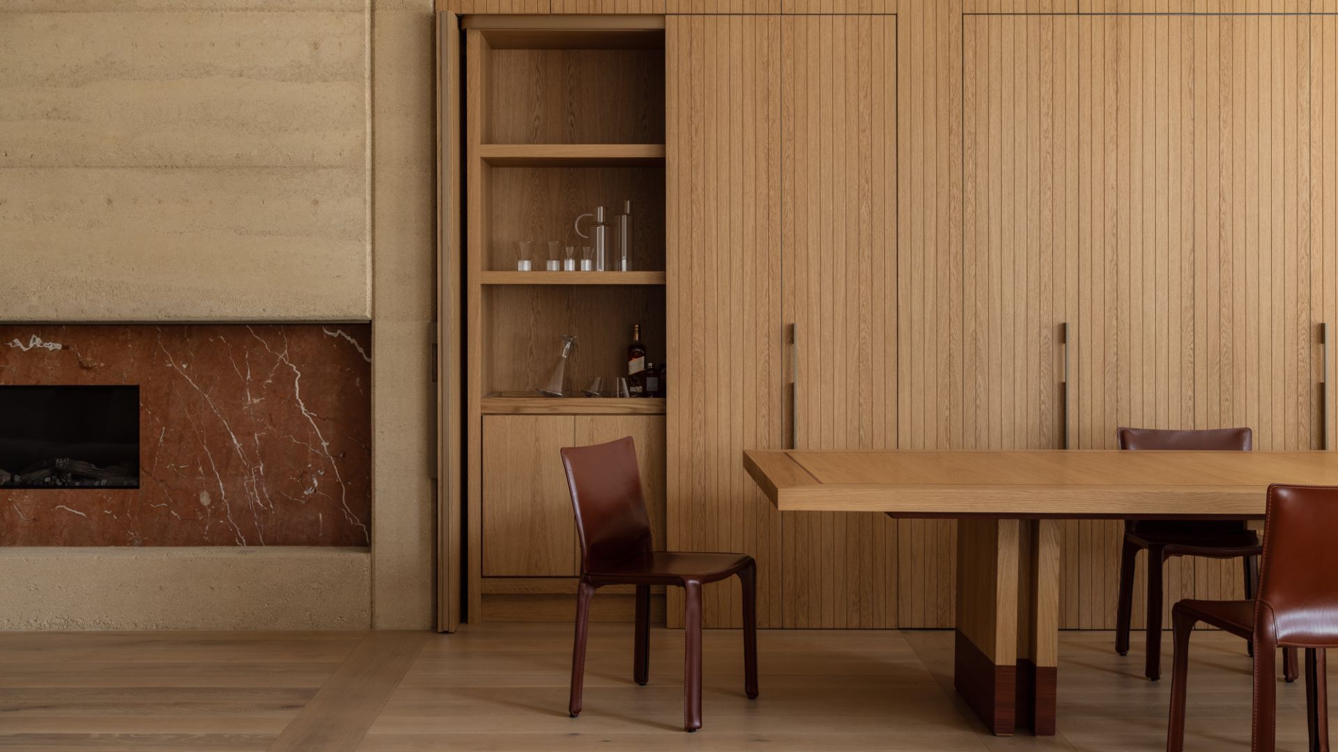 A minimalist dining room with wood cupboards a long wood table, shiny leather seating, and a stone fireplace
