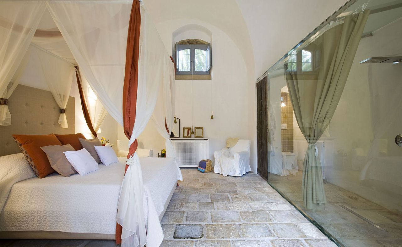 A hotel room with a four post bed, various colour cushions, a white chair, a white console, square stone flooring and a tinted glass wall between the bathroom.