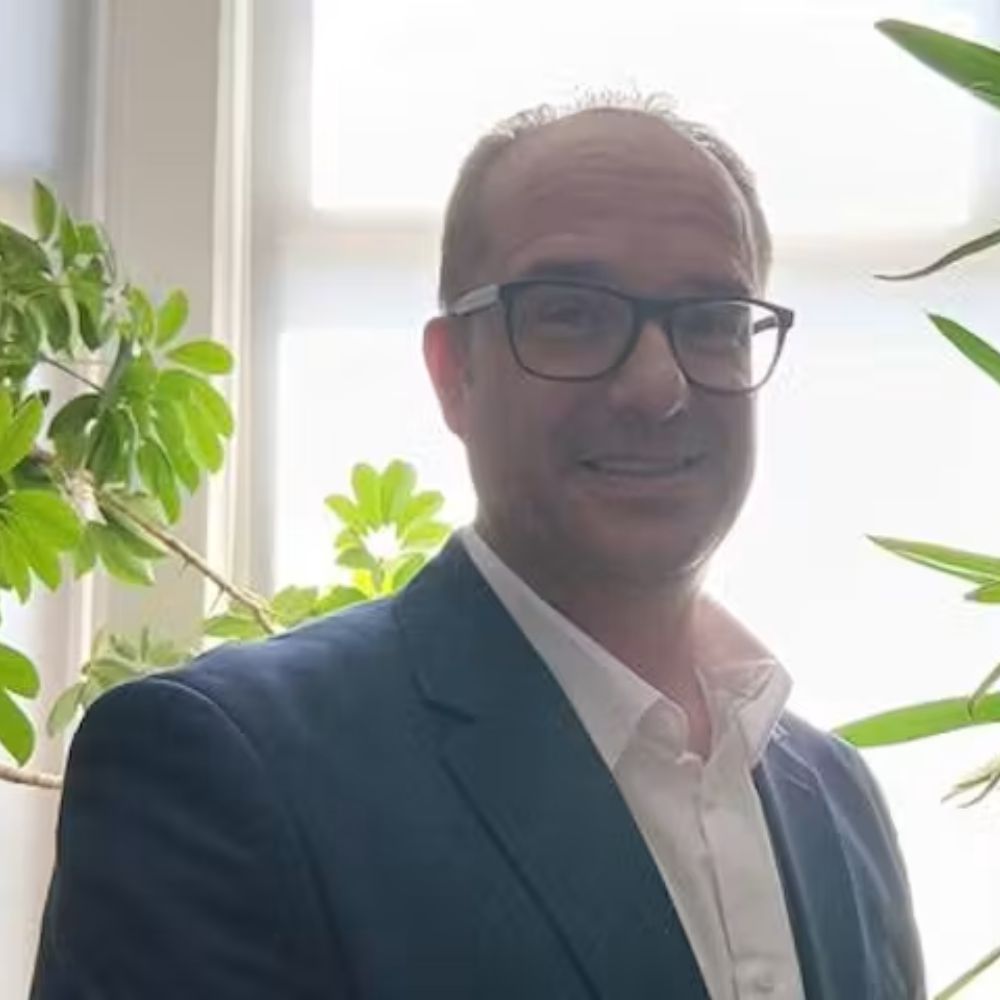A headshot of a man in a suit smiling at the camera surrounded by plants