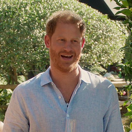 Prince Harry has red hair and beard and wears a light blue linen shirt while holding a champagne glass in With Love, Meghan