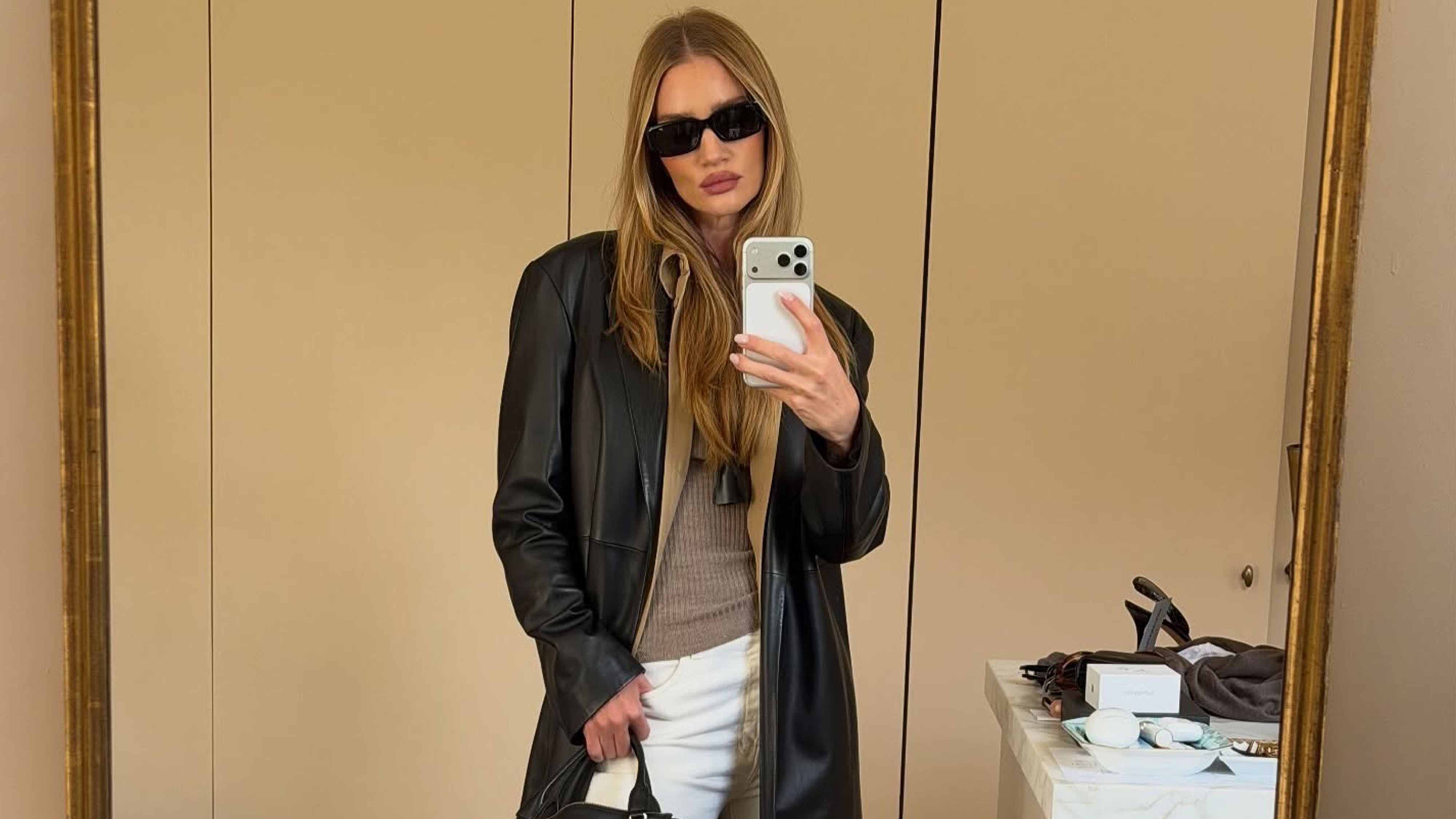 Rosie wears sunglasses, a long black leather coat, pendant necklace, and white jeans.