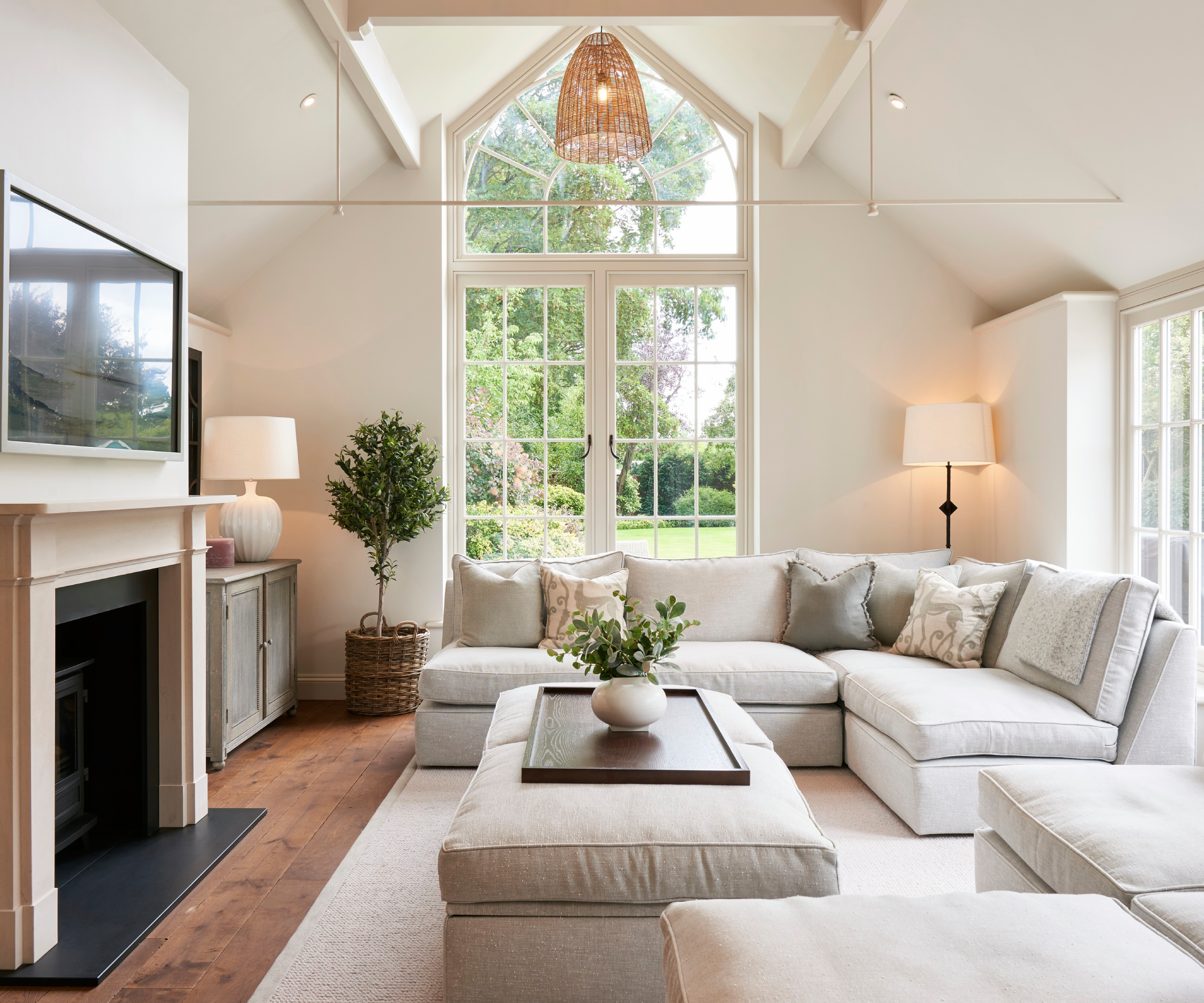 living room with large arched window at end, cream sofas and coffee table placed facing fireplace with TV above, patio doors behind, large cream rug and wooden flooring
