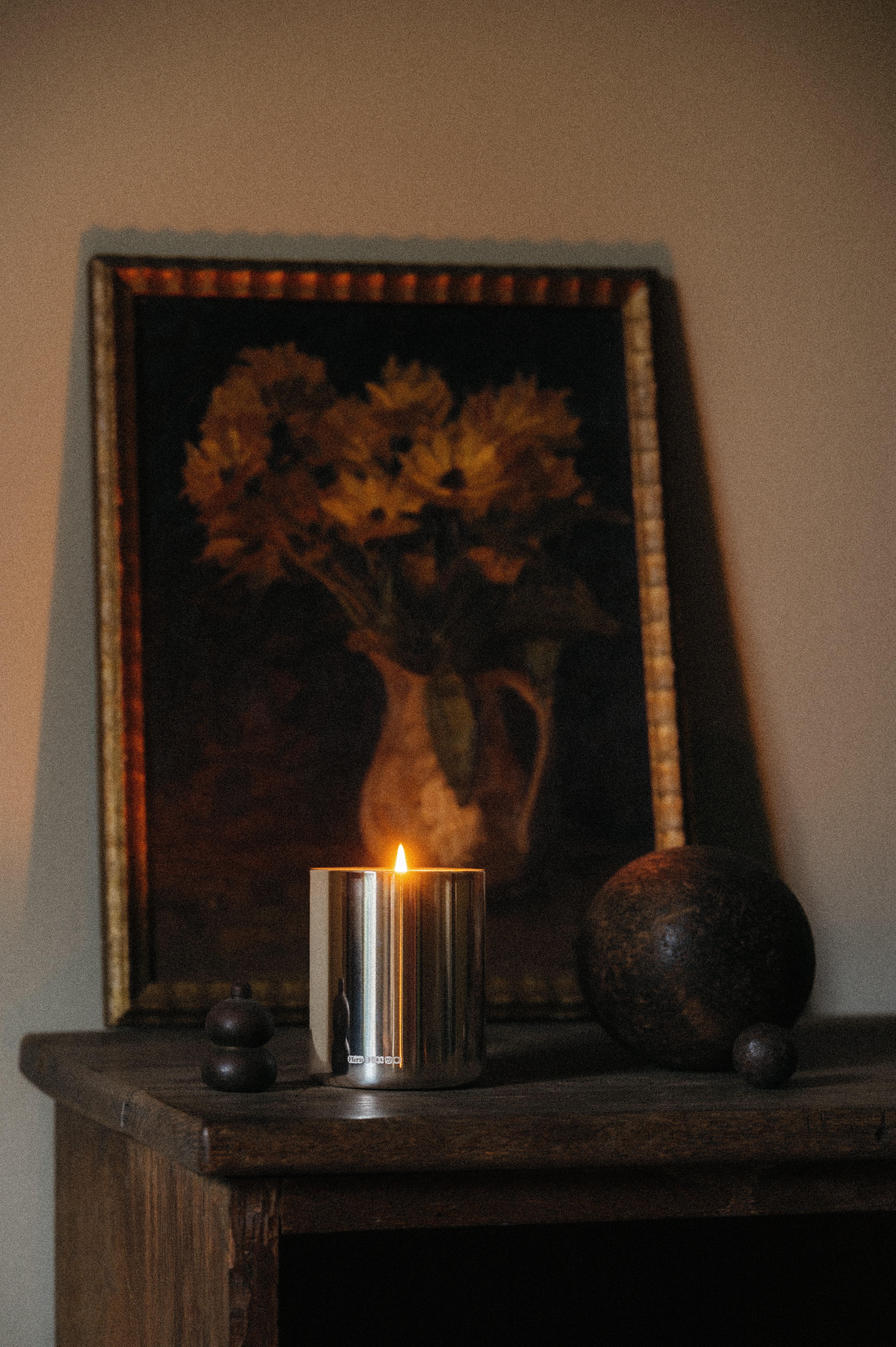 A chrome-packaged candle burns atop a wooden chest of drawers in front of a floral painting framed in gold and next to two abstract wooden sculptures.