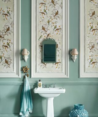 A bathroom with white wall panels with brown bird wallpaper, teal walls, two white wall sconces, a white standing basin, and a light blue towel