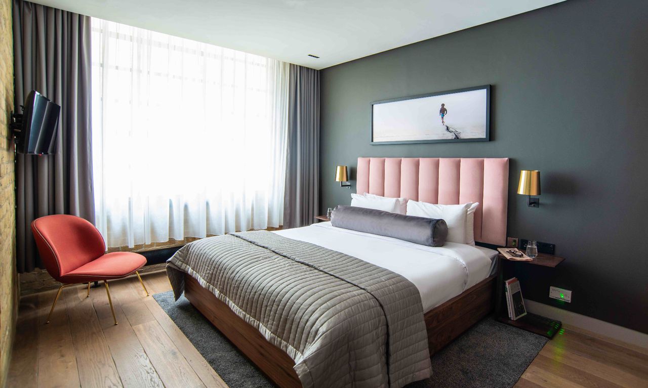 Interior view of a bedroom at one of Native&#039;s ApartHotels featuring a wooden bed with pink headboard, wall-mounted television, chair and large window