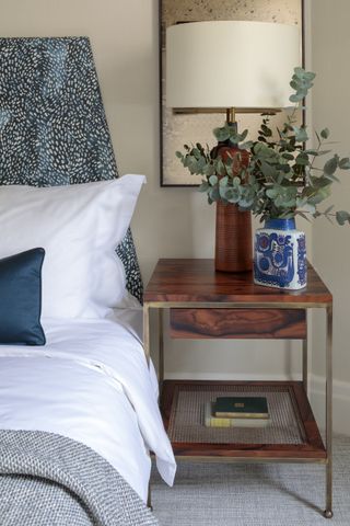 small bedroom with mahogany style side table, gray carpet, large table lamp with mahogany coloured base, vase, blue headboard and cushion, white bedding, gray throw
