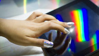 a person with light skin and sparkly long nails uses a touchscreen with a rainbow reflected on it