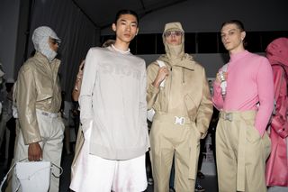 Three models - one with a pale grey sweater with DIOR print, one in a camel-coloured outfit, and one in a pink shirt with camel-coloured trousers