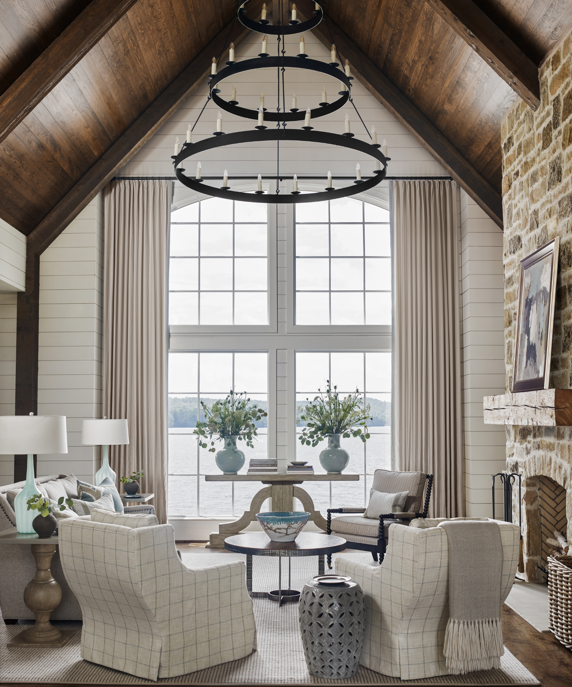 Lake house living room featuring two slipcovered accent chairs, sky blue details, a brick fireplace, two-tier rustic chandelier, and tall windows overlooking the water.