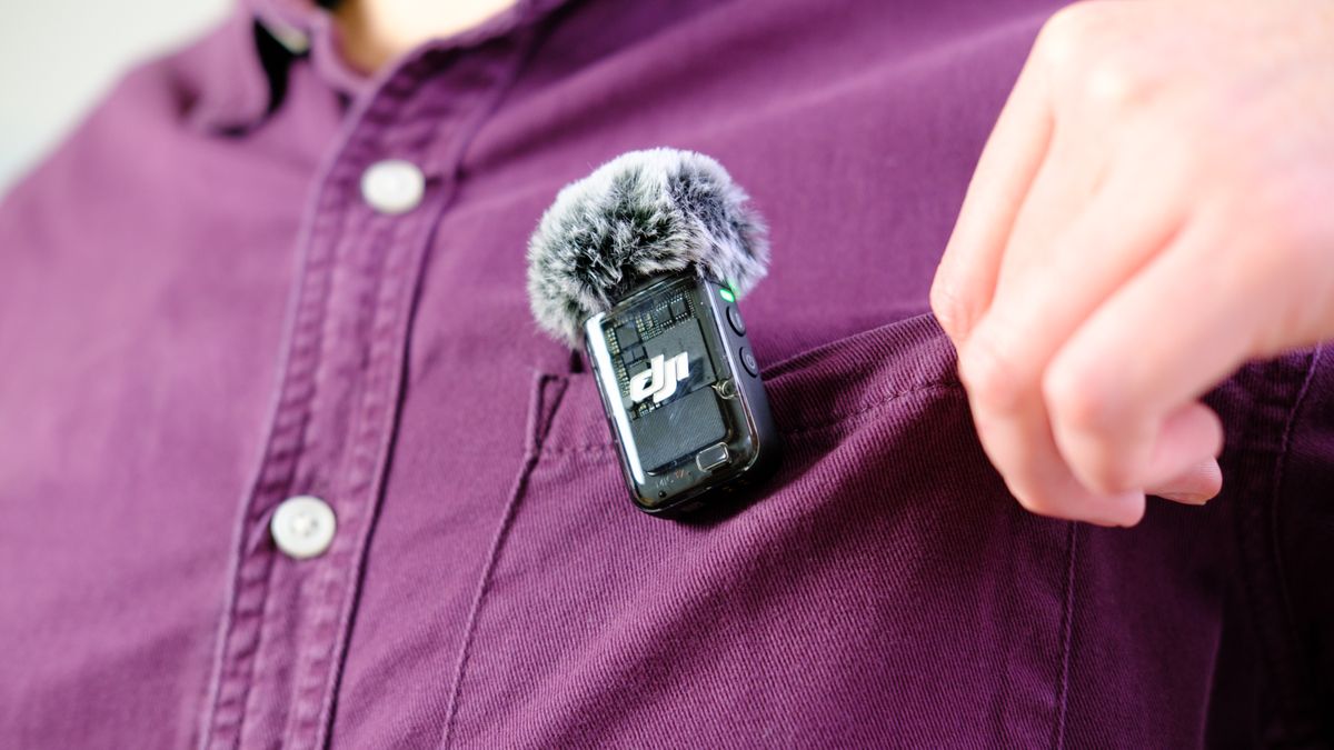 A photo of the DJI MIc 2 TX unit attached to the pocket of a burgundy shirt — the wearer of the shirt is adjusting the pocket for comfort. The mic is fitted with a wind shield.