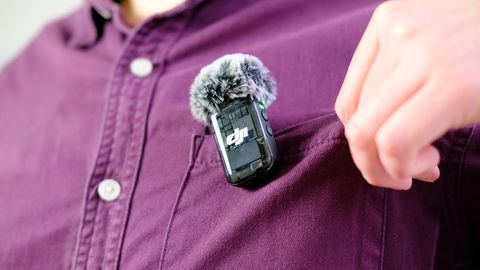 A photo of the DJI MIc 2 TX unit attached to the pocket of a burgundy shirt — the wearer of the shirt is adjusting the pocket for comfort. The mic is fitted with a wind shield.