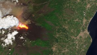 A satellite photo shows a glowing lava river flowing down the side of Mount Etna as a plume of ash and gas is released from its other side