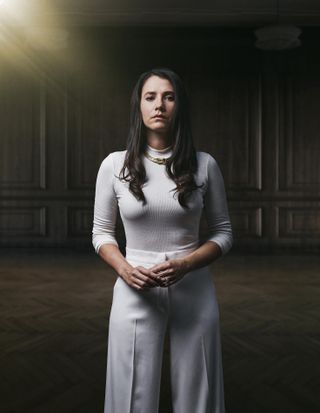 A posed shot of Natalie Varga (Charlie Murphy), dressed in a white long-sleeved top and white trousers, standing in a panelled room, looking into the camera as she toys with the wedding ring on her finger