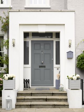 freshly rendered stylish grey front door at the top of white steps, with a welly and umbrella stand at the door at flowers planted in grey pots at the foot of the stairwell
