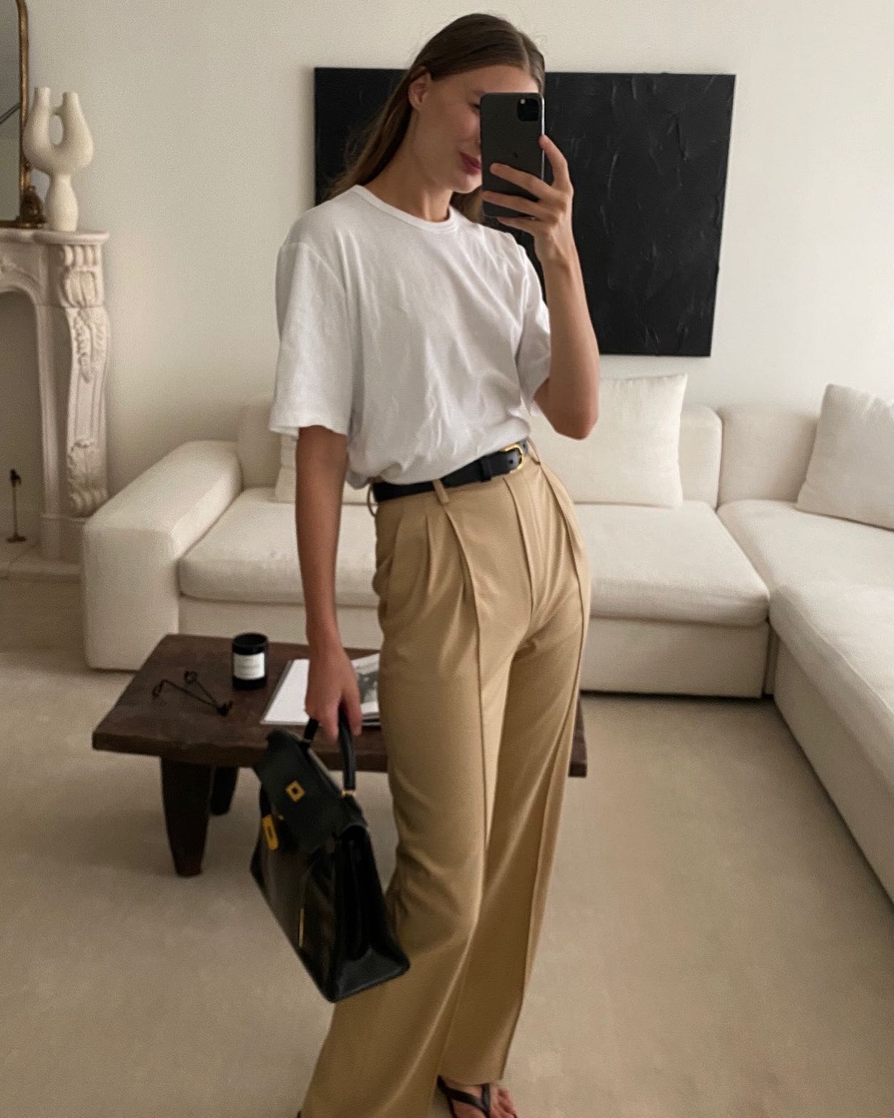 fashion influencer Christie Tyler poses for a mirror selfie wearing a white t-shirt, thin black belt, Hermes bag, tan pleated pants, and black flip-flop sandals