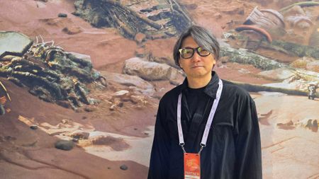 Gen Atlas creator Fumito Ueda standing in front of promotional art for the game.