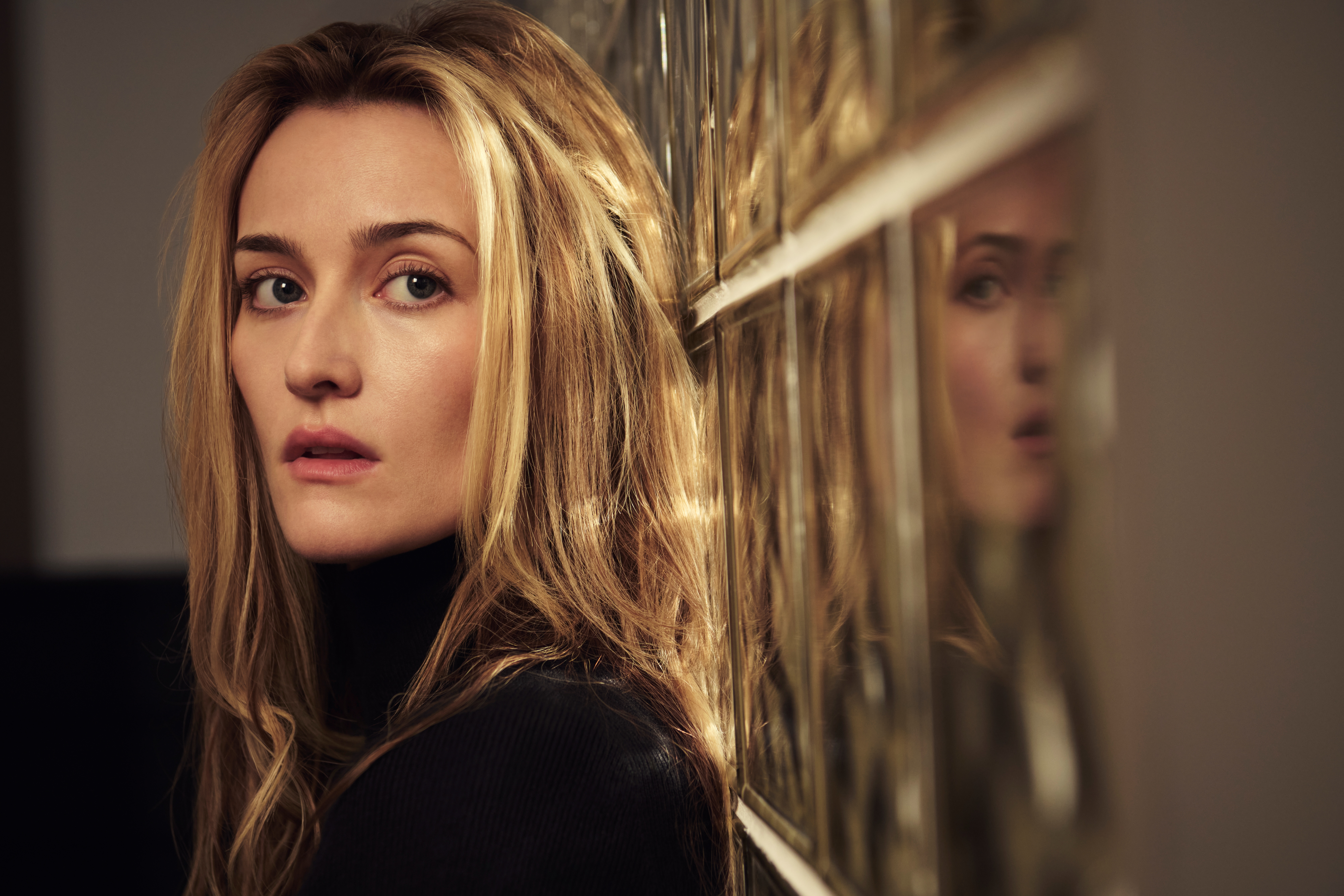 A young blonde woman wearing a black turtleneck stands with her head resting against a glass brick wall.