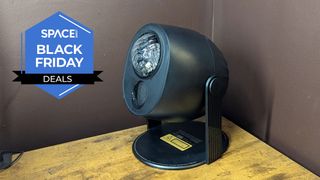 A black projector is on top of a wooden surface with a dark colored wall in the background.