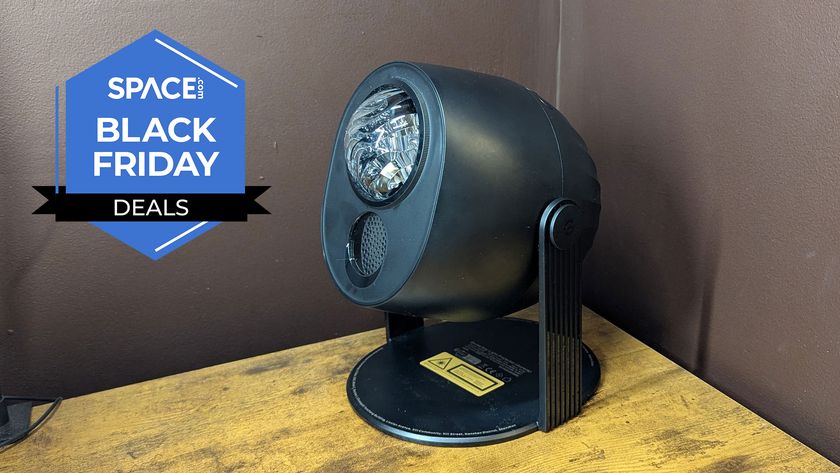 A black projector is on top of a wooden surface with a dark colored wall in the background.