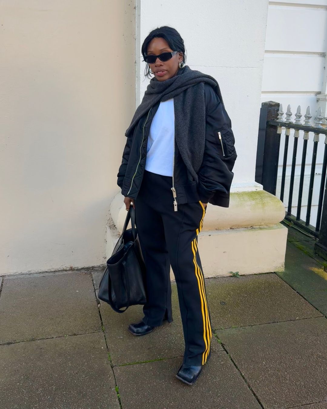 @anuogugbesan wears a black bomber jacket with a white t-shirt, Adidas joggers with yellow side stripes and black boots whilst standing in the street