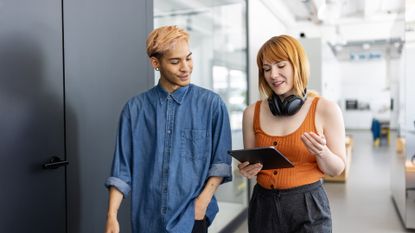 Male and female Gen Z tech workers walking through an open plan office space while holding a tablet computer. 