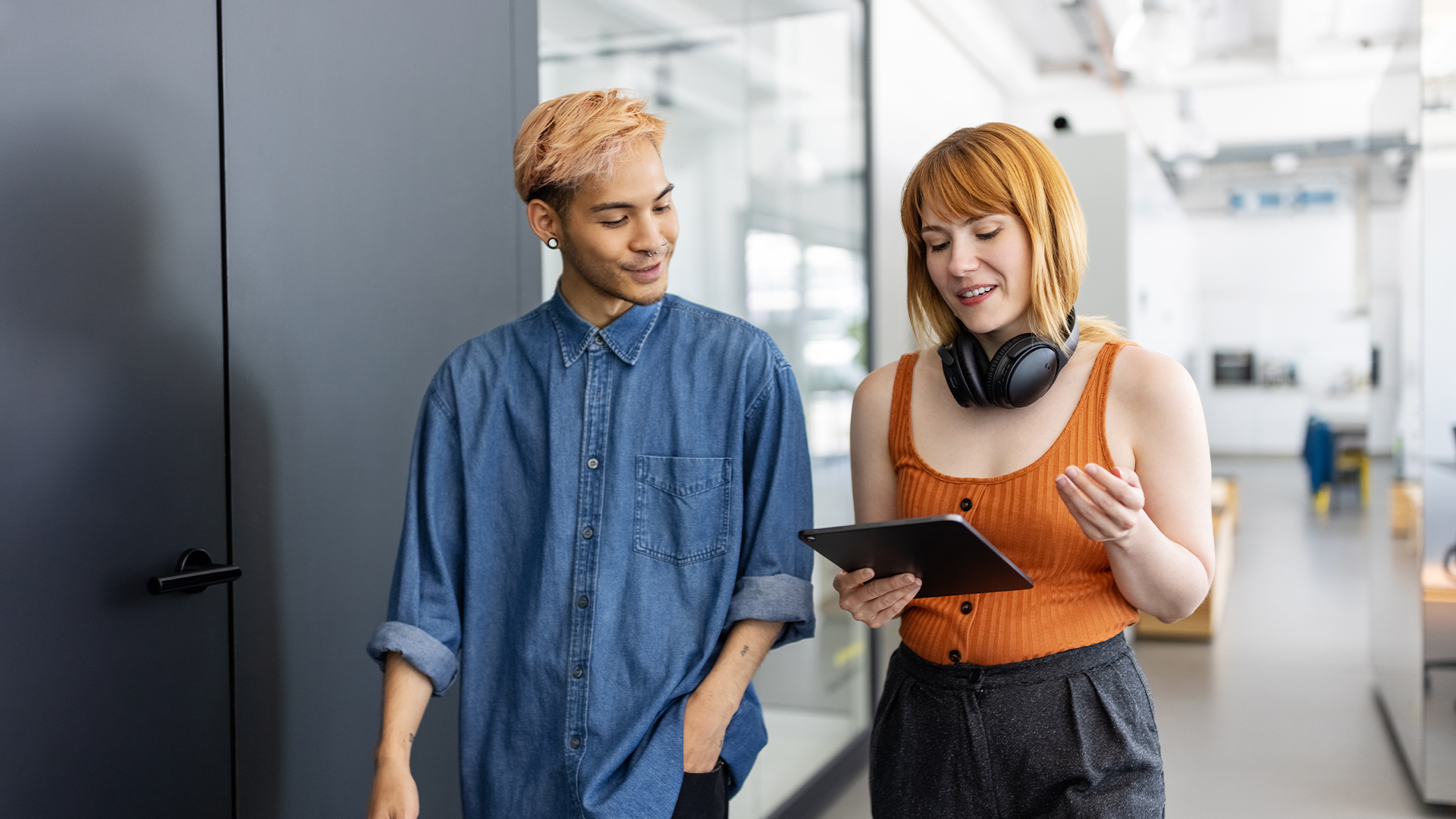 Male and female Gen Z tech workers walking through an open plan office space while holding a tablet computer. 