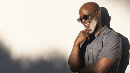 An older man wearing sunglasses smiles as he strokes his beard, appearing deep in thought.
