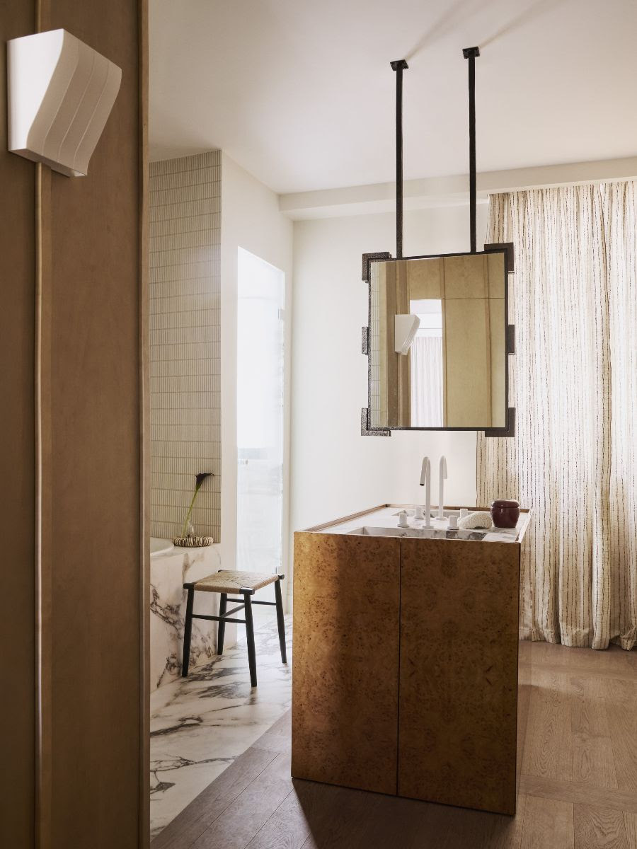 a modern bathroom with timber floor and marble floor around bath, curtains, tiled wall, timber panelling, and a central vanity in the room wrapped in burl veneer with a suspended mirror hanging above
