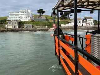 Alan Carr stands on the deck of the 'sea tractor' that transports guests to and from Burgh Island when the tide is in. The Art Deco style Burgh Island hotel is behind him on the island.