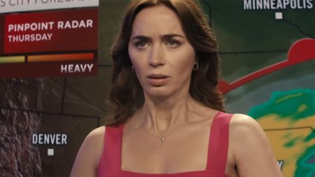 Emily Blunt sits behind a weather desk in a pink dress looking scared in Disclosure Day. 