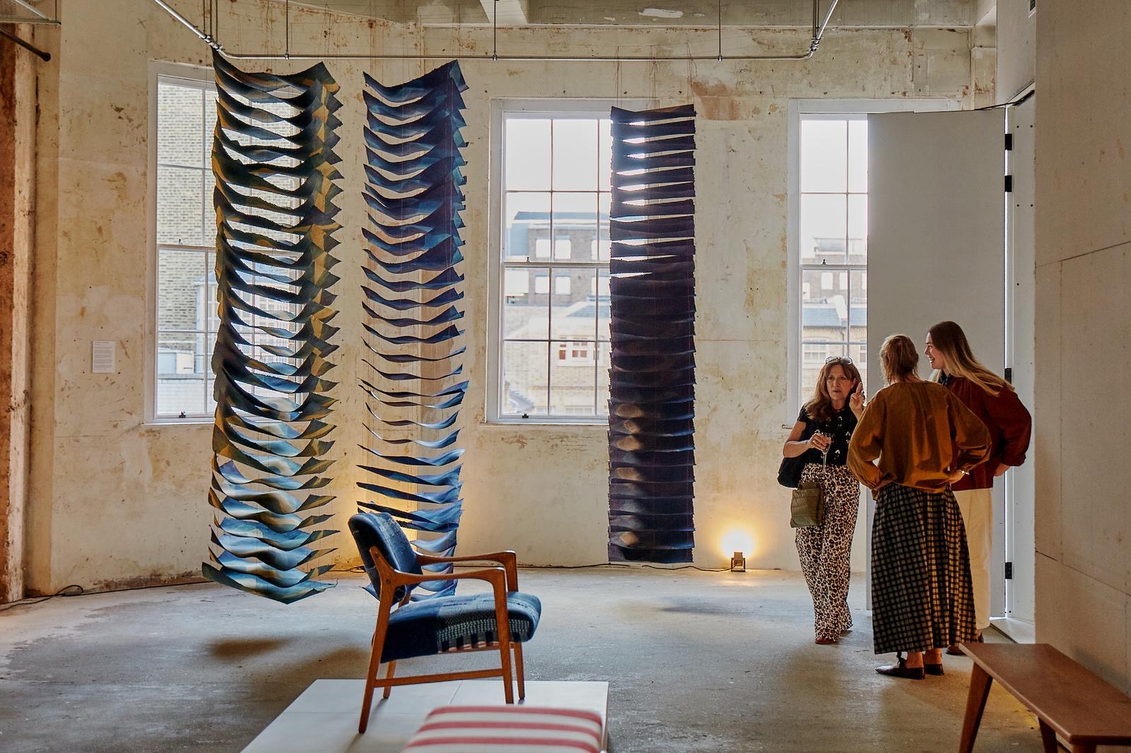 A series of installation shots from a show hosted inside a historical building with unfinished walls and tall ceilings captures the soft atmosphere inside it, punctuated by floating artworks, crafty furniture and accessories, and people chatting or performing.