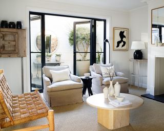 monochrome living room with two linen armchairs, 1970s wooden coffee table, rattan chair and black metal framed doors