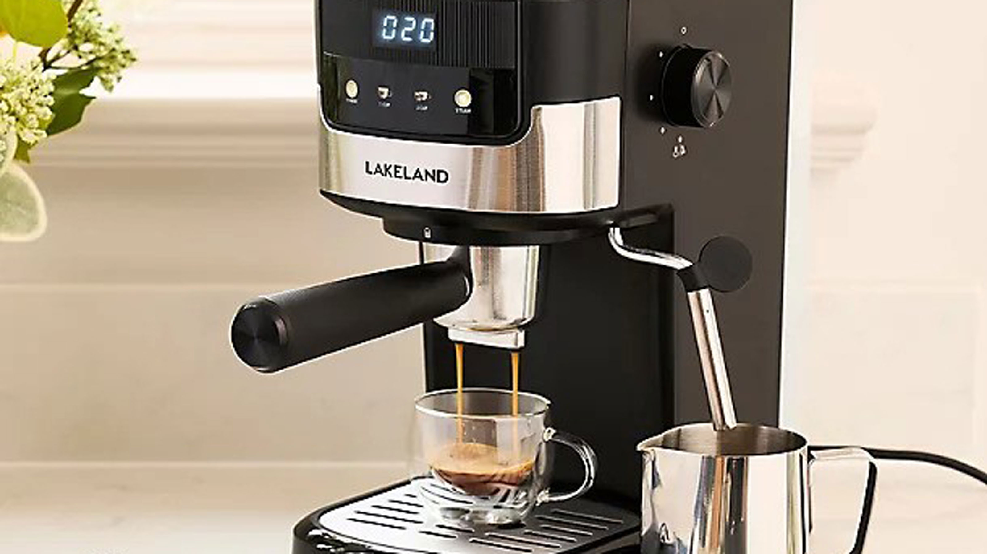 Black coffee machine with portafilter pulling a shot of espresso over a clear cup and a steam wand in a chrome milk jug