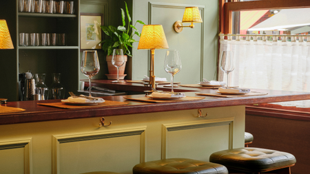 A sage green restaurant with a wood-top bar set with placemats, plates, cutlery, and wine glasses