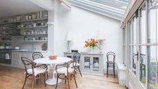 Conservatory with glass doors, round white breakfast table, wooden bistro chairs, a side console with two large lamps, leading into a kitchen with lots of open shelving decorated with ceramics