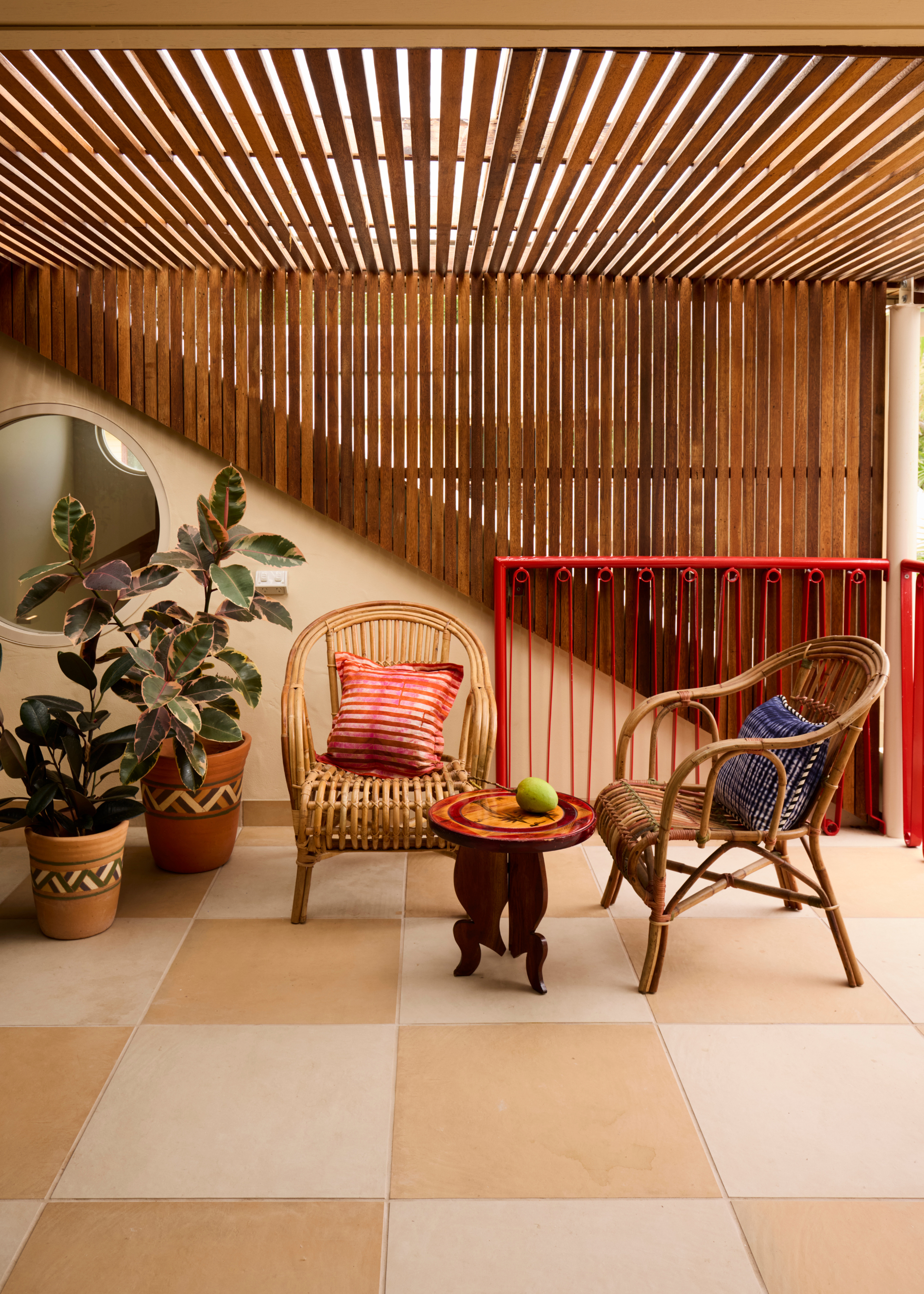 Vertical timber battens cover a slatted ceiling on a balcony&amp;rsquo;s lounge area, which go down the wall in a triangular shape, with two wicker chairs in front and two plant pots, with a porthole window beside