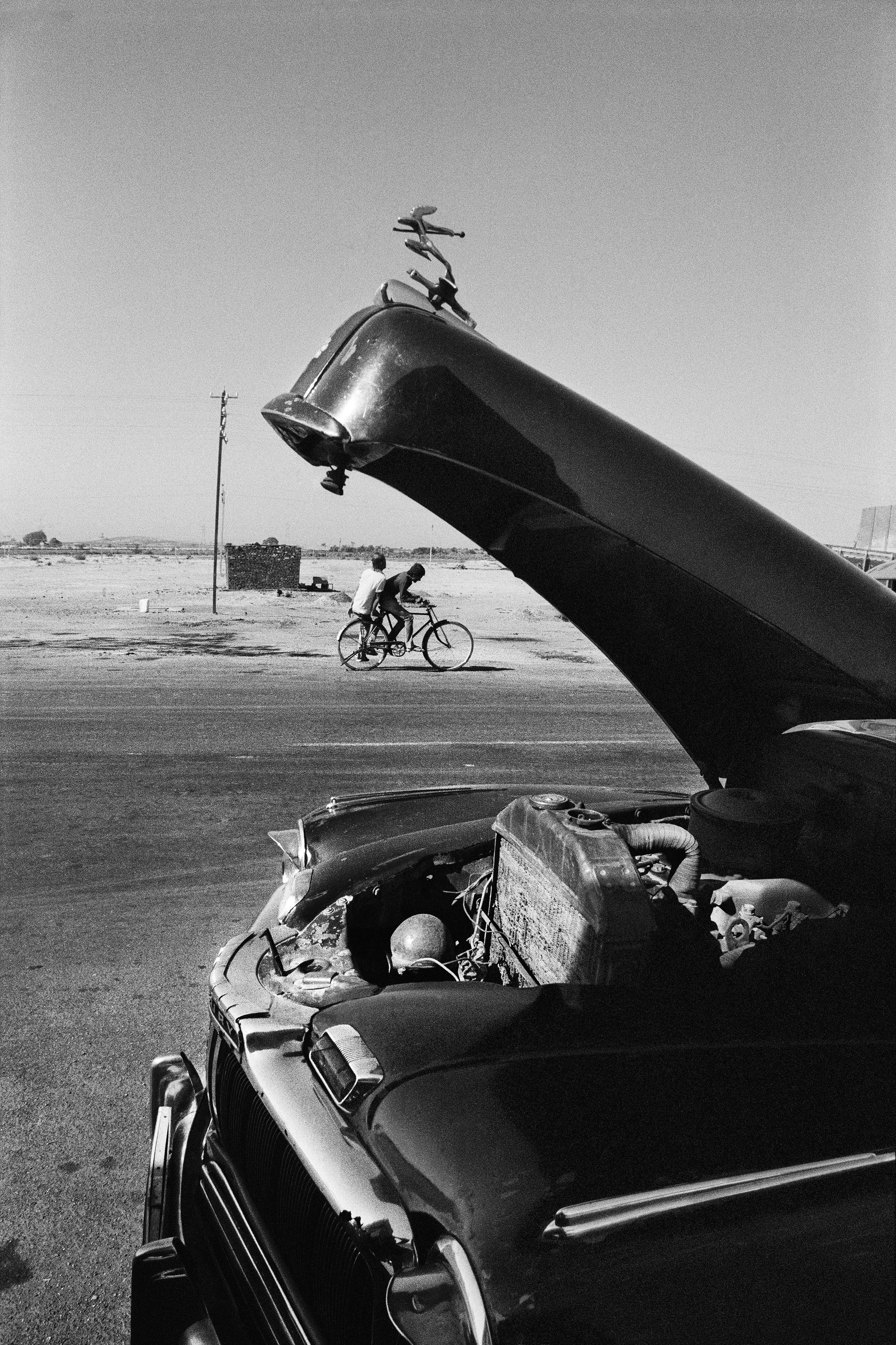 A vintage car with its hood open is parked by the roadside, while two cyclists ride past in the background under a clear sky