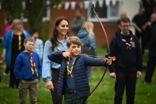 Prince George shooting a bow and arrow with Princess Kate standing behind him on grass