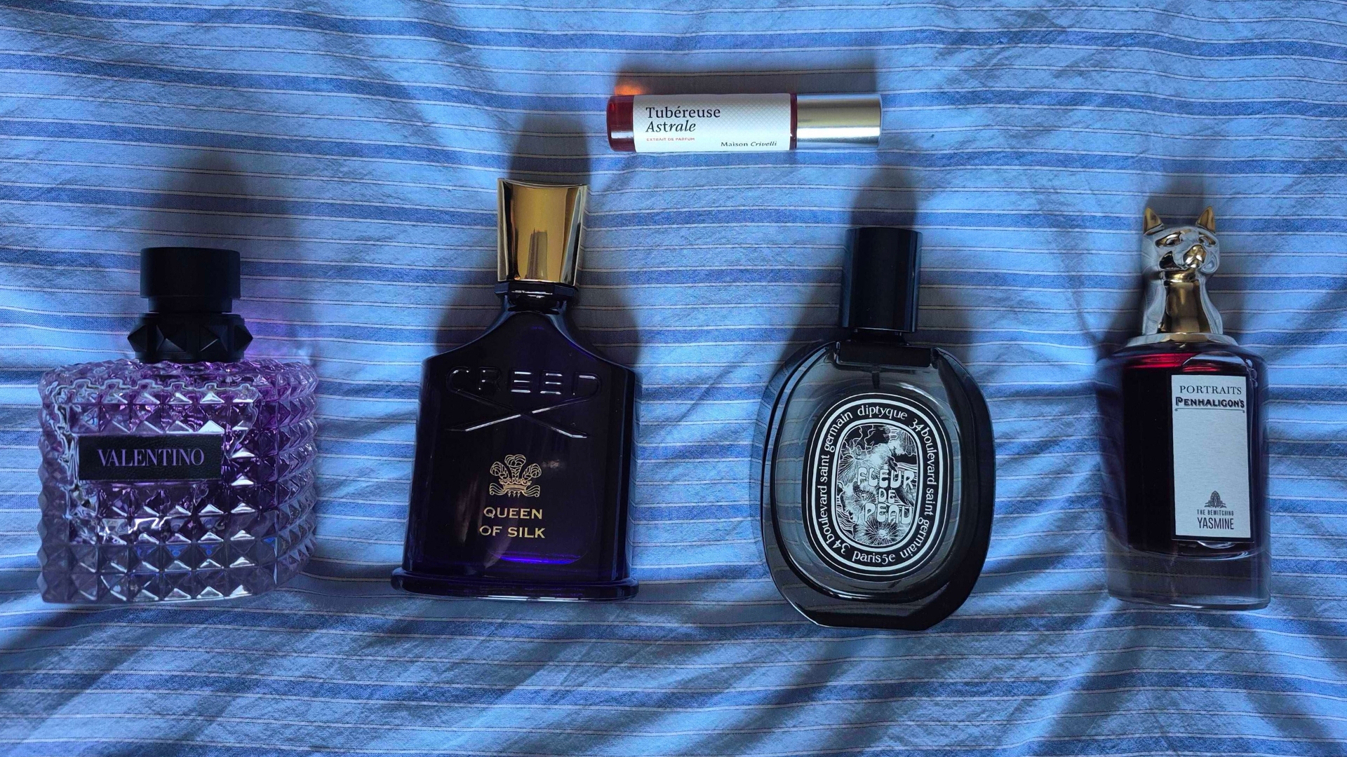 A collection of some of the perfumes included in this Evening Florals Feature, including the Valentino Roma Melancholia Donna parfum, Creed's Queen of Silk, Diptyque's Fleur de Peau, Penhaligon's The Bewitching Yasmine and Maison Crivelli's Tubereuse Astrale, all laid out on a blue and white-striped bedspread.