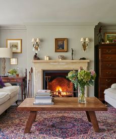 An eclectic living room with a large marble lit fireplace, patterned purple rug, light matching sofas with assorted pillows, a large wooden coffee table with stacks of books and a vase of large flowers, a dark brown chest of drawers, framed artworks and sage green walls