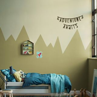 Sage green wall with painted triangles, blue bed with patterned bedspread, sotrage cabinet and wall hanging