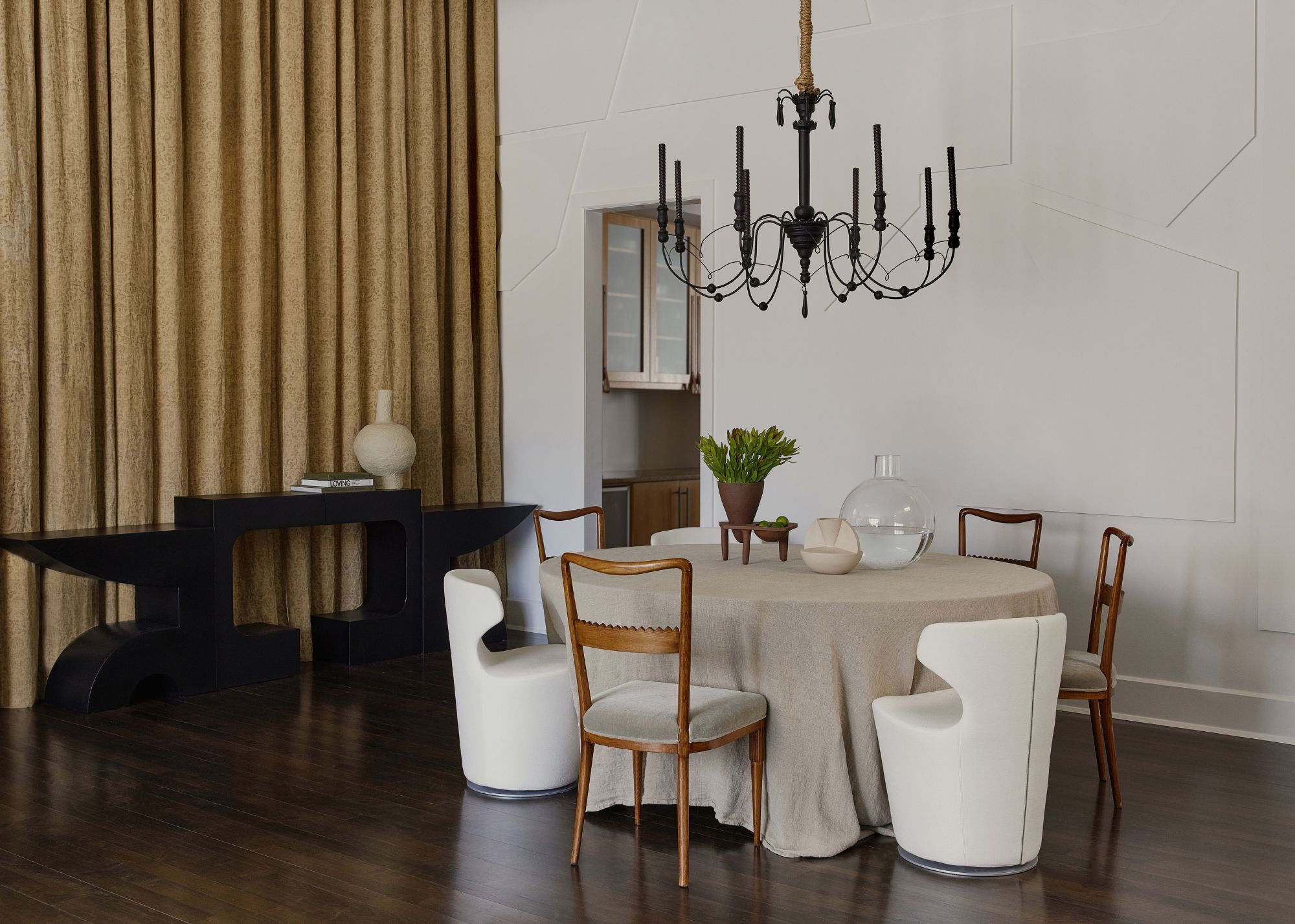 A dining room with dark wood flooring, a black painted chandelier, four vintage chairs and two armchairs, and a black console