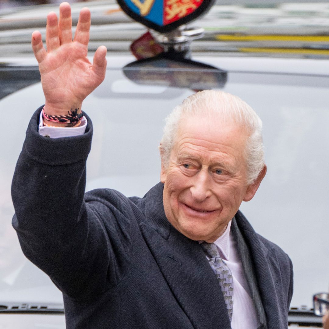 King Charles waving wearing a gray coat in front of a car